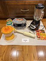 Three kitchen appliances displayed on white surface: yellow and white Salton juicer, white and gray Braun hand blender, gray and black Black & Decker electric grill with lid and accessories nearby.