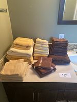 Seven stacks of bathroom towels in beige, yellow, and brown on a bathroom counter near a sink and mirror.