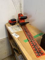 Electric hedge trimmer with long dual blade on a wooden table with battery and instruction manual nearby.