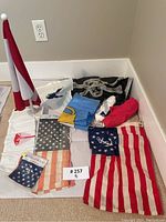 Overview image of all flags in the lot spread on floor including a plastic red and white mini flag on a white base, several folded fabric flags including American and other designs.