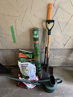 Photo of garden shovel, grass seed bag, mesh netting roll, soil/fertilizer bags, and watering can against a concrete wall.
