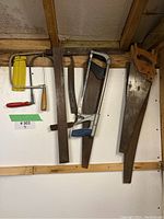 Full view of hand saws and wood tools hanging on wall showing 2 hacksaws, a bow saw, a try square, and a coping saw