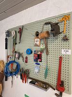 Lot of assorted hand tools arranged on a green pegboard, including pliers, mallet, hand saw, pipe wrench, cutters, measuring tape, blue tape, and other small hand tools and accessories