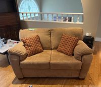 Front view of tan microfiber loveseat with two patterned throw pillows on hardwood floor near white railing.