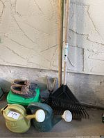 Photo showing green Miracle Gro plastic tub plant food and assorted gardening tools including wooden rakes, watering cans, hand gardening tool, and gardening shoes on concrete floor by wall.