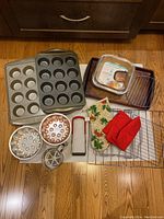 Overview of bakeware items including muffin pans, bundt pans, baking dishes, oven mitts, cooling racks, and sifter on a wood floor.
