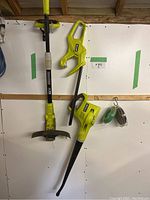 Ryobi brand string trimmer and leaf blower hung on white pegboard with green tape.