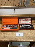 Two socket sets side by side on a workbench, one in an orange metal case and one in a red metal case, showing included ratchets and multiple sockets.