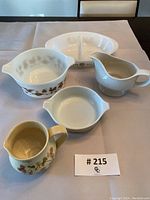 Five vintage kitchenware items arranged on a table with a white background: divided white serving dish, bowl with brown design, gravy boat, small white dish with handles, and floral pitcher.