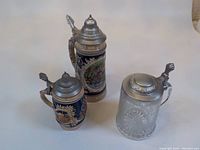 Three beer steins together showing tall Heidelberg stein, smaller ceramic stein, and glass stein with pewter lids and decorated exteriors.