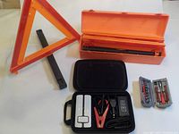 Photo of orange box containing three expanding reflective road triangles with foldable legs, a black zippered case with jump starter accessories, and a small gray tool case with screwdriver bits visible.
