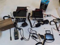 Photo showing 4 black CB radio units with cables, microphones, Baofeng handheld radio, Nextar GPS, boxed antenna, power cords laid out on table