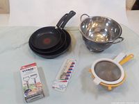 Photo showing three black T-Fal frying pans stacked by size, stainless steel colander, two strainers with yellow and white handles, Titan peeler box, and a packaged set of colorful measuring spoons.