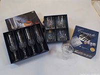 Photo showing the two boxed sets: the Concerto whiskey tumblers box with four short crystal glasses and the Longchamp Cristal d'Arques champagne flutes box with four tall cut crystal flutes, one tumbler out of box for viewing.