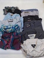 Group photo of 12 men's button down shirts folded and stacked. Colors and patterns include plaid, solid blue, striped, and neutral colors.