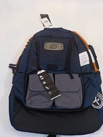 Front view of the Naruto Shippuden kids backpack showing navy blue and orange color scheme, metal ninja symbol plate, two gray flap pockets, and tags attached.