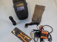 Photo of lot with welding helmet, gloves, flashlight, stick holder, crimp tool, and battery charger.
