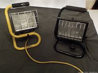 Two portable halogen bulb work lights, one with a yellow frame and one with a black frame, both with protective grilles and padded handles.