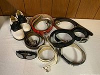 Photo of white and black ladies golf shoes, seven belts in various colors including black, brown, red, white perforated, and tan, and a pair of black sunglasses with brown lenses on a white surface.