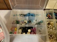 Clear plastic organizer box with compartments filled with various beads, pendants, and found jewelry parts including blue, white, pink, and black beads.