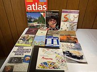 Photo showing 15 books arranged on a white surface with wood panel background, including travel atlas, art book, cooking books, and photography basics.