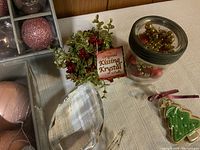 Close-up of green and red faux berry sprig labeled Kissing Krystal, pink ball ornaments in box, glass candle holders, glass jar with red and gold ornaments, and ceramic Christmas tree cookie ornament.