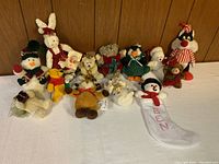 Group photo showing all plush toys spread out on table in front of wood panel wall including snowmen, teddy bears, Winnie the Pooh, and Christmas stocking.