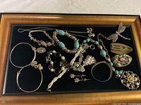 Full view of assorted costume jewelry including bracelets, earrings, brooches, and pins with a black velvet background and a gold-tone wooden frame