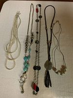 Five costume jewelry necklaces laid out on a beige surface showing the different styles, bead colors, and length of each necklace.