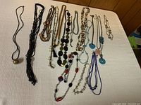 Photo of 14 assorted costume jewelry necklaces laid out on white fabric surface, showing variety in styles, materials, and colors.