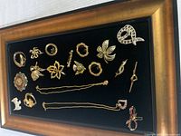 Framed black velvet tray display showing multiple costume jewelry pieces including brooches and pins in various shapes and finishes, mostly gold-tone with some rhinestones and pearls.