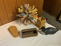 Six decorative wooden and natural material items displayed on white cloth against wood-paneled background: driftwood ball, pen holder, bird figurine, carved box, wooden block and tray.