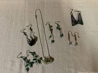 Photo of five pairs of earrings and one necklace with green stone pendant on silver chain displayed on light fabric surface.