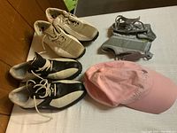 Photo showing two pairs of ladies golf shoes, two gray wrist supports, and a pink Wilson baseball cap laid out on a surface.