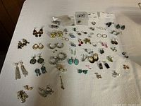 Wide shot showing the majority of the earrings spread on a beige fabric surface, highlighting different sizes, colors, and styles.