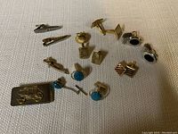 Multiple men's cuff links and tie clips spread out on a beige fabric surface showing design details and quantity.