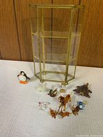 Photo showing a brass framed glass display case and 10 miniature animal figurines, including a penguin, turtle, camels, ducks, and others on a white surface with a wood background