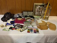 Overview of lot including framed painting, glass lantern, wooden plaques, boxed craft items, hat, tangled cables, and a silver electronic device on a white table