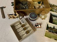 Photo showing wooden box filled with assorted jewelry, small figurine, decorative container, cocktail picks in original packaging, wooden horse figurine, metal corkscrew opener, and six vintage postcards arranged on table.