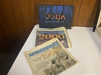Photo showing an Outsiders Ouija Board game box and two vintage newspapers: one from 1900 and another from 2000, both from Toronto Star publications.