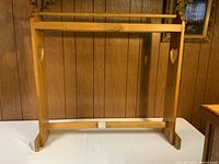 Wooden quilt rack featuring heart-shaped cutouts on vertical sides, two horizontal bars for hanging quilts or blankets, light wood finish, placed on a table with wood-paneled wall background.