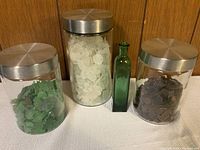 Photo showing three glass jars filled with sea glass pieces in white, dark green, and dark brown colors with silver metal lids, alongside one small green glass bottle.