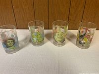 Four clear drinking glasses each decorated with different Shrek characters laid out on a white surface against a wooden background.