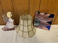 Photo showing the stained glass angel figurine, a geometric beige lamp shade, and a square stained glass floral suncatcher leaning against a wooden background.