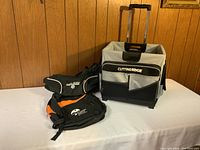 Gray and black Cutting Edge rolling tote with handle and two front pockets alongside two backpacks, one black and white Cutting Edge, and one black and orange Sporting Goods