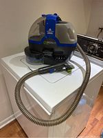 Front and side view of the Bissell SpotBot Pet carpet cleaner on a white washer or dryer showing the hose and cleaning tool.