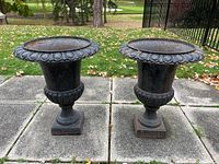 Front view of two cast iron urns on concrete patio, showing decorative rims and pedestal bases