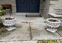 Photo showing both cement urns placed on a concrete walkway near a front porch with a dark door and stone wall background.