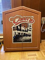 Front view of closed wooden dartboard cabinet with Cheers artwork