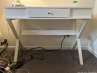 White desk with one drawer and angled legs, showing front view with drawer closed and plugged-in lamp on top.